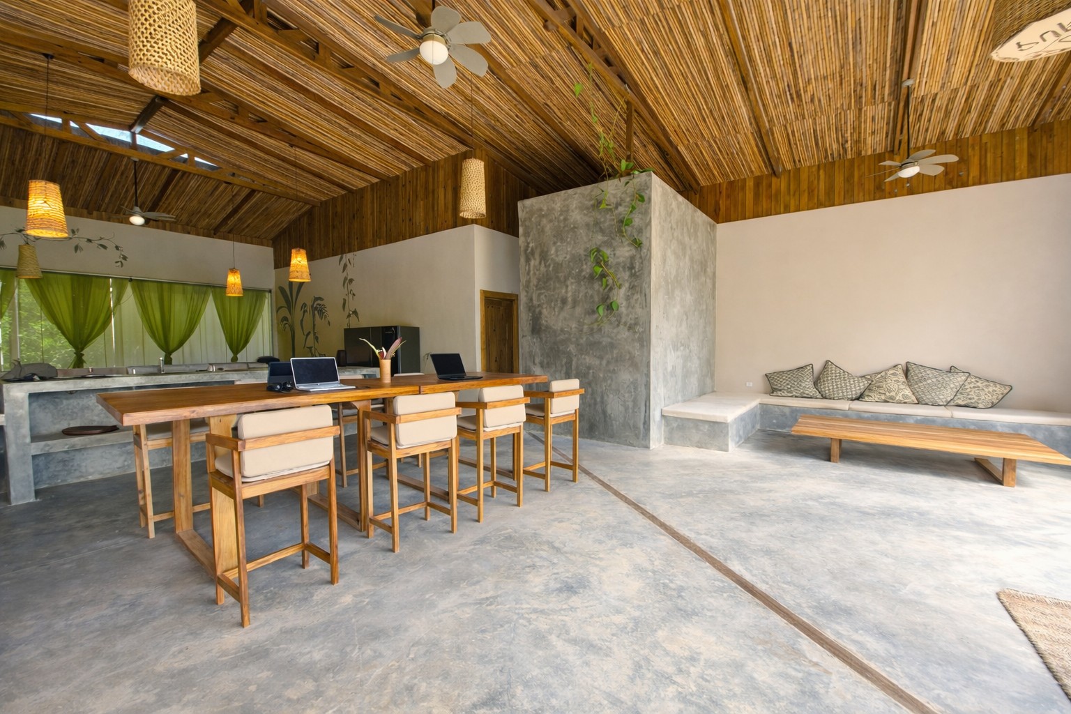 Mesa alta de madera con taburetes y, al lado, un rincón de sofá con cojines, bajo el techo de caña